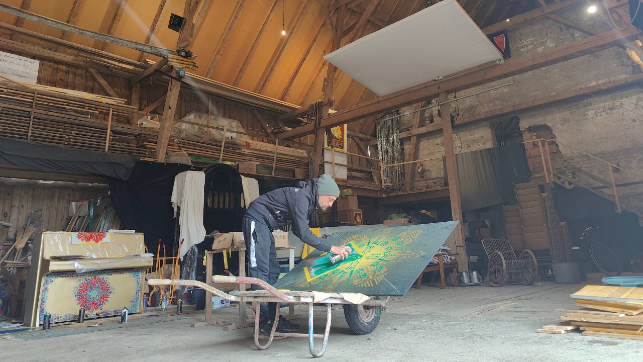 Der Künstler Erwa.One aus dem Streetart / urbane Kunst gestaltet ein Holzpaneel in einer Scheune, welches an ein Klavier (links im Bild) angebracht wird). Er ist bei der Arbeit und besprüht die Holztafel mit seinen Schablonen aus dem Design "Neue Ornamente"