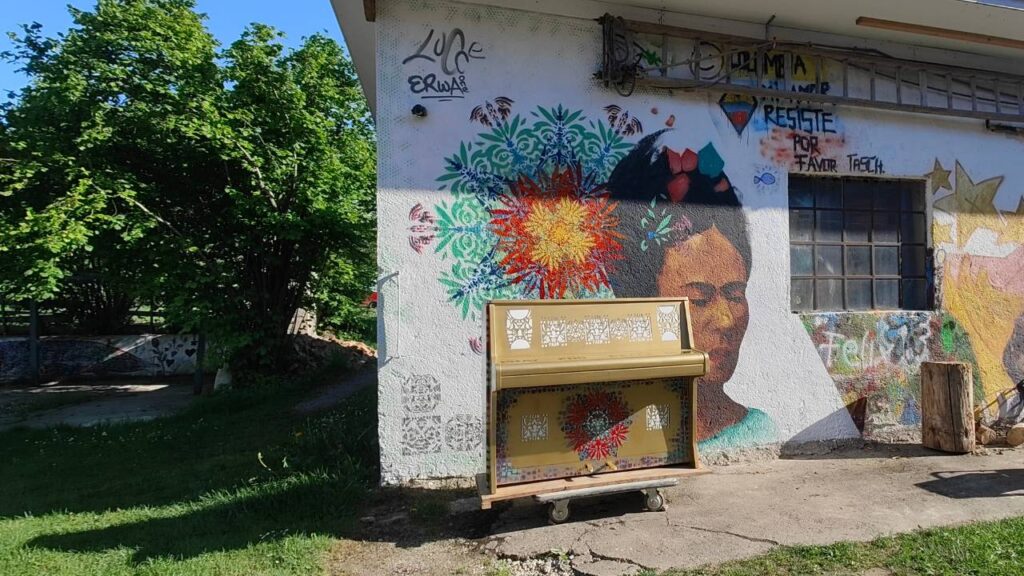 Play Me Klavier gestaltung Pray for Peace Play durch Erwa.One. Das Klavier steht vor einem Streetartwerk und ist mit bunten Ornamenten geschmückt auf goldenem Hintergrund.