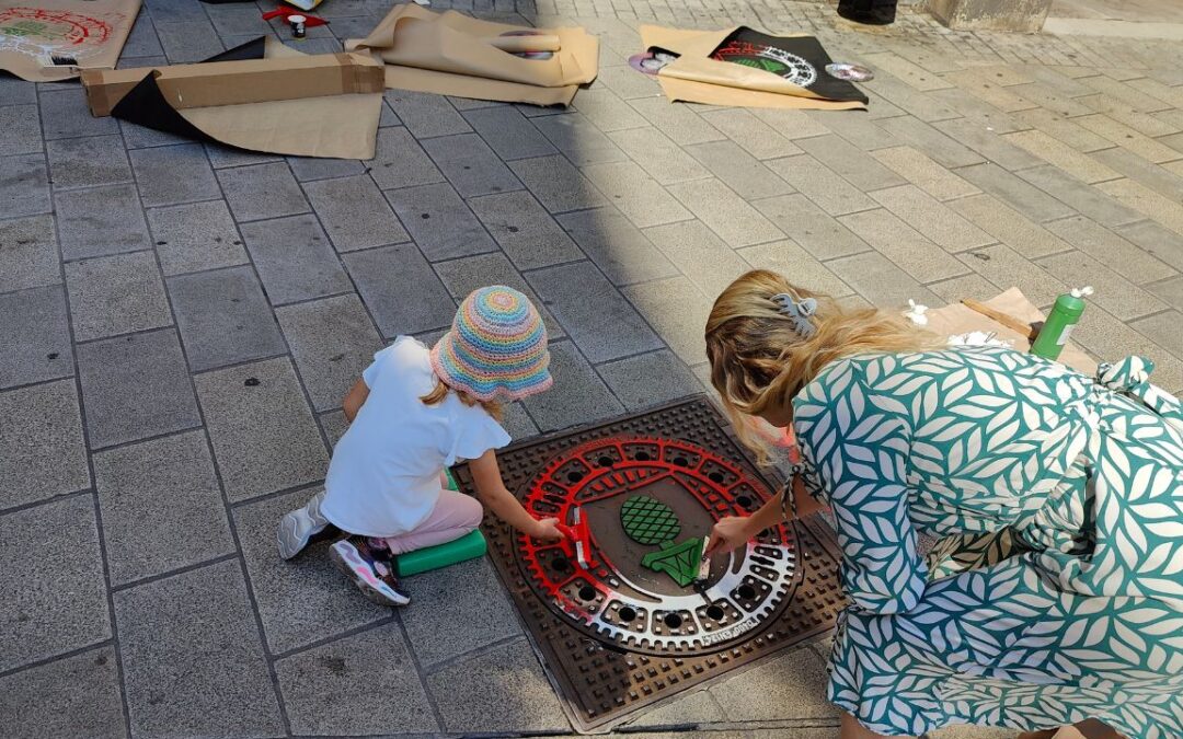 Print the City Streetart Intervention durch ErwaOne in Augsburg in Kooperation mit den Kunstsammlungen und museen von Augsburg vor dem Maximiliansmuseum (Fuggerplatz). Ein kleines Mädchen walzt mit ihrer Mutter Farbe auf einen Gulli-Deckel