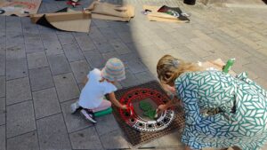 Print the City Streetart Intervention durch ErwaOne in Augsburg in Kooperation mit den Kunstsammlungen und museen von Augsburg vor dem Maximiliansmuseum (Fuggerplatz). Ein kleines Mädchen walzt mit ihrer Mutter Farbe auf einen Gulli-Deckel