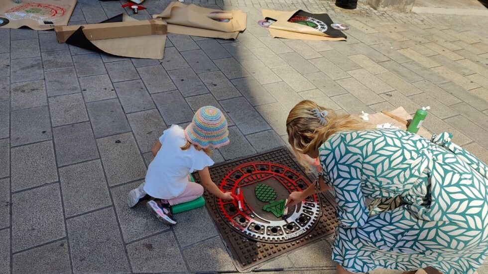 Print the City Streetart Intervention durch ErwaOne in Augsburg in Kooperation mit den Kunstsammlungen und museen von Augsburg vor dem Maximiliansmuseum (Fuggerplatz). Ein kleines Mädchen walzt mit ihrer Mutter Farbe auf einen Gulli-Deckel