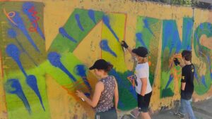 Intergenerationeller Streetart- und Graffiti Workshop mit ErwaOne in Landsberg am Lech an der legalen Sprühfläche der Stadt. Workshophelferin Emma sprüht neben einem Kind ein Graffito mit jeweils den Buchstaben K und U für Kunst. Grüner Hintergrund