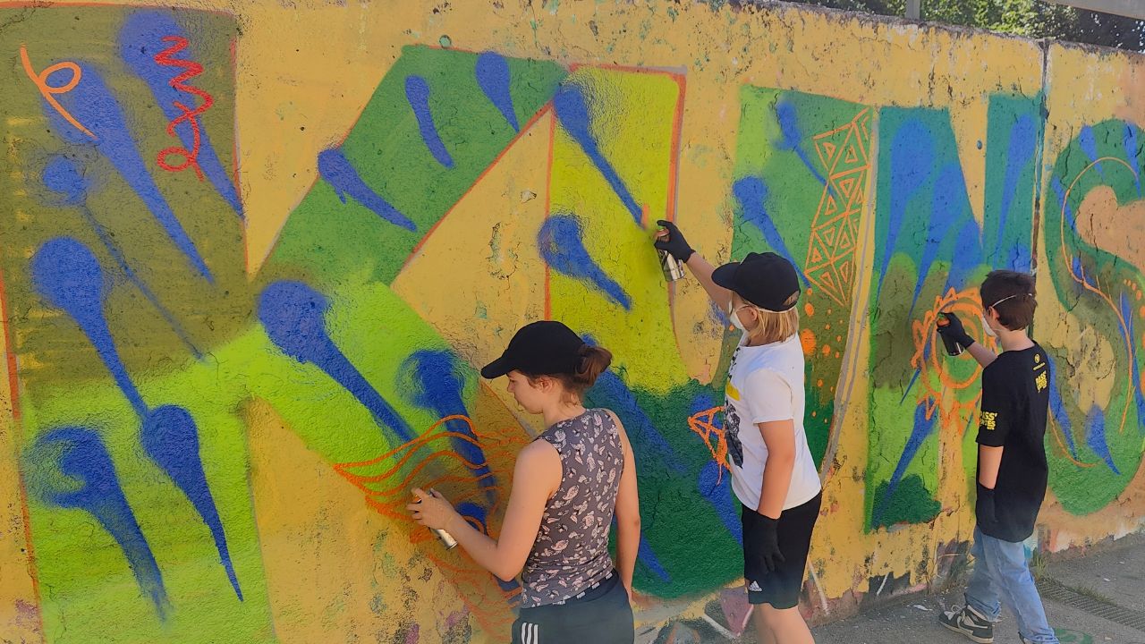 Intergenerationeller Streetart- und Graffiti Workshop mit ErwaOne in Landsberg am Lech an der legalen Sprühfläche der Stadt. Workshophelferin Emma sprüht neben einem Kind ein Graffito mit jeweils den Buchstaben K und U für Kunst. Grüner Hintergrund