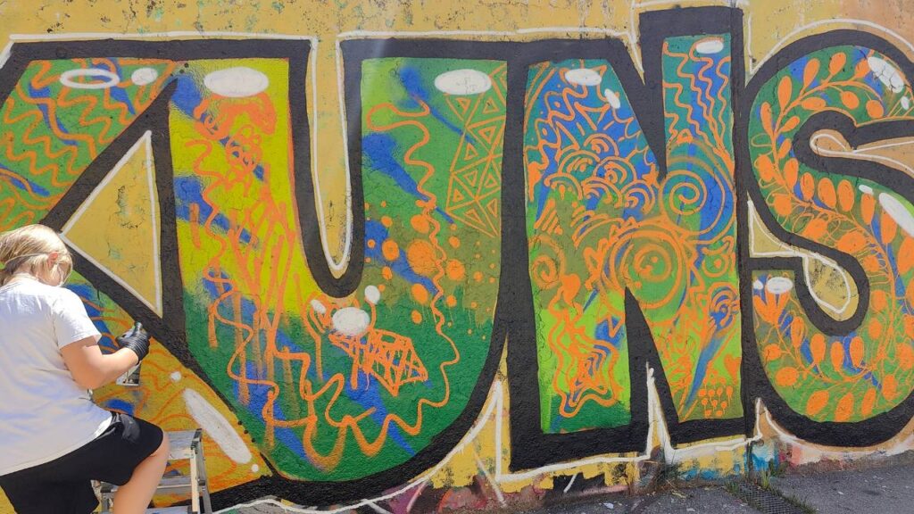 Intergenerationeller Streetart- und Graffiti Workshop mit ErwaOne in Landsberg am Lech an der legalen Sprühfläche der Stadt. Ein Junge sprüht einen Buchstaben und malt die innere Fläche aus. Das Wort Kunst ist zu lesen