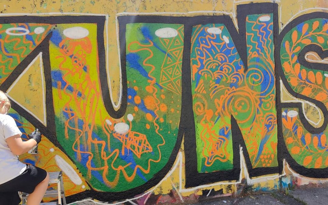 Intergenerationeller Streetart- und Graffiti Workshop mit ErwaOne in Landsberg am Lech an der legalen Sprühfläche der Stadt. Ein Junge sprüht einen Buchstaben und malt die innere Fläche aus. Das Wort Kunst ist zu lesen