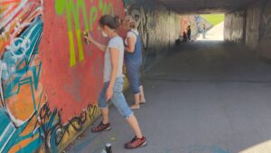 Intergenerationeller Streetart- und Graffiti Workshop mit ErwaOne in Landsberg am Lech an der legalen Sprühfläche der Stadt. Zwei Mütter sprüht ein Graffito mit dem Schriftzug Mémé für ihre Großmutter, welche bald Geburtstag hat und 90 Jahre alt wird