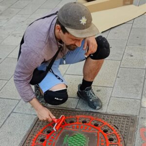 Print the City Streetart Intervention durch ErwaOne in Augsburg in Kooperation mit den Kunstsammlungen und museen von Augsburg vor dem Maximiliansmuseum (Fuggerplatz). Der Künstler walzt Farbe auf einen Gulli Deckel mit dem Motiv der Zirbelnuss -Augsburgs Wahrzeichen