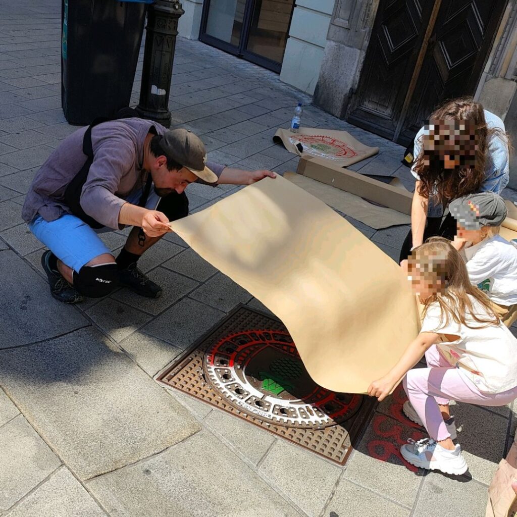 Print the City Streetart Intervention durch ErwaOne in Augsburg in Kooperation mit den Kunstsammlungen und museen von Augsburg vor dem Maximiliansmuseum (Fuggerplatz). Eine Familie legt mit dem Künstler ErwaOne ein Papier auf einen Gulli Deckel, um diesen abzudrucken.