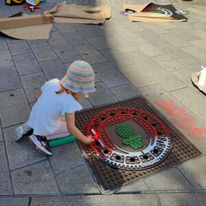 Print the City Streetart Intervention durch ErwaOne in Augsburg in Kooperation mit den Kunstsammlungen und museen von Augsburg vor dem Maximiliansmuseum (Fuggerplatz). Ein kleines Mädchen walzt Farbe auf einen Gulli-Deckel