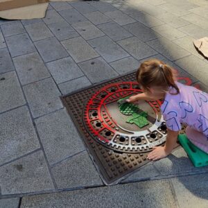 Print the City Streetart Intervention durch ErwaOne in Augsburg in Kooperation mit den Kunstsammlungen und museen von Augsburg vor dem Maximiliansmuseum (Fuggerplatz). Ein kleines Mädchen walzt Farbe auf einen Gulli-Deckel