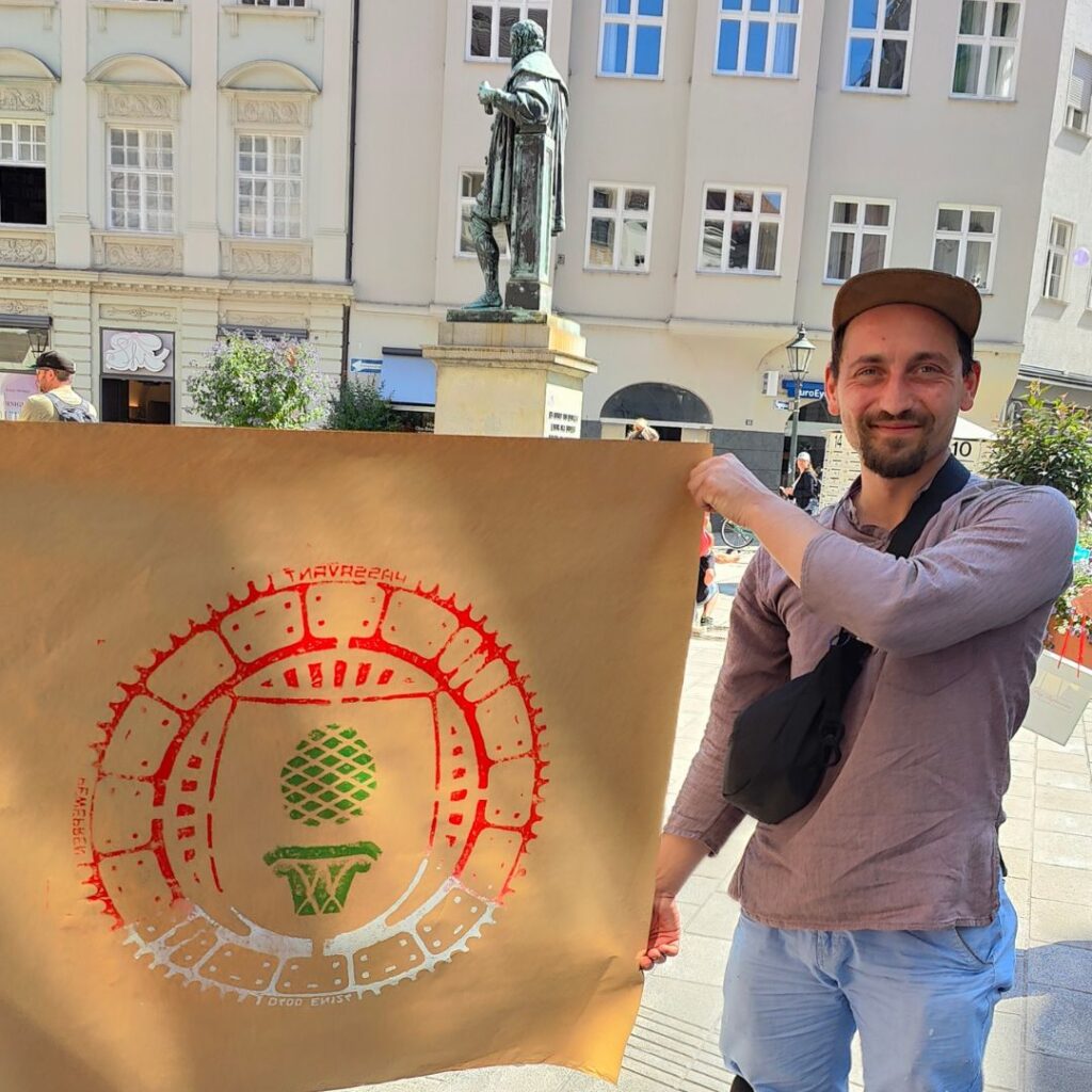 Print the City Streetart Intervention durch ErwaOne in Augsburg in Kooperation mit den Kunstsammlungen und museen von Augsburg vor dem Maximiliansmuseum (Fuggerplatz). EDer Künstler hält einen Gulli-Deckel Abdruck in die Kamera. Hinter im sieht man eine Statue
