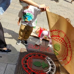 Print the City Streetart Intervention durch ErwaOne in Augsburg in Kooperation mit den Kunstsammlungen und museen von Augsburg vor dem Maximiliansmuseum (Fuggerplatz). Eine Familie legt mit dem Künstler ErwaOne machen einen Gulli-Deckel Abdruck