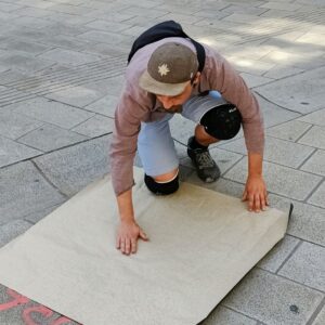 Print the City Streetart Intervention durch ErwaOne in Augsburg in Kooperation mit den Kunstsammlungen und museen von Augsburg vor dem Maximiliansmuseum (Fuggerplatz). Der Künstler drückt ein Papier auf einen Gulli-Deckel