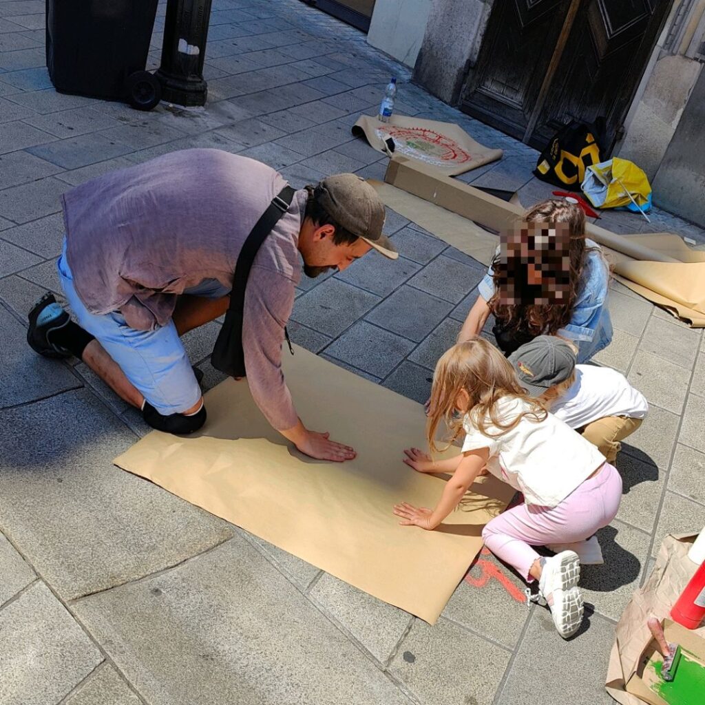 Print the City Streetart Intervention durch ErwaOne in Augsburg in Kooperation mit den Kunstsammlungen und museen von Augsburg vor dem Maximiliansmuseum (Fuggerplatz). Der Künstler drückt ein Papier auf einen Gulli-Deckel. Zwei Kinder und eine Mutter helfen mit den Abdruck der Zirbelnuss zu drucken.