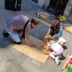 Print the City Streetart Intervention durch ErwaOne in Augsburg in Kooperation mit den Kunstsammlungen und museen von Augsburg vor dem Maximiliansmuseum (Fuggerplatz). Der Künstler drückt ein Papier auf einen Gulli-Deckel. Zwei Kinder und eine Mutter helfen mit den Abdruck der Zirbelnuss zu drucken.