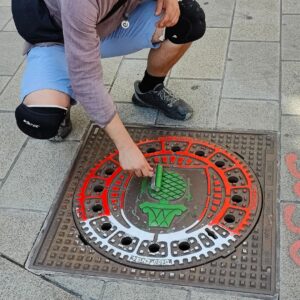 Print the City Streetart Intervention durch ErwaOne in Augsburg in Kooperation mit den Kunstsammlungen und museen von Augsburg vor dem Maximiliansmuseum (Fuggerplatz). Der Künstler reibt den Gulli Deckel mit dem Zirbelnuss Motiv mit einer Farbwalze in den Augsburger Farben ein grün weiß rot