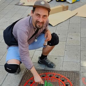 Print the City Streetart Intervention durch ErwaOne in Augsburg in Kooperation mit den Kunstsammlungen und museen von Augsburg vor dem Maximiliansmuseum (Fuggerplatz). Der Künstler reibt einen Gulli Deckel mit einer Farbwalze ein und schaut in die Kamera