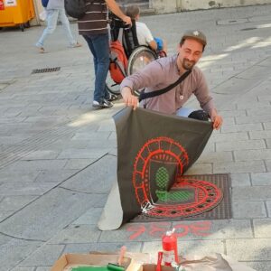 Künstler ErwaOne druckt einen Gulli Deckel mit dem Motiv der Zirbelnuss am Fuggerplatz vor dem Maximiliansmuseum von Augsburg ab