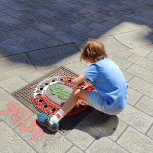 Print the City Streetart Intervention durch ErwaOne in Augsburg in Kooperation mit den Kunstsammlungen und museen von Augsburg vor dem Maximiliansmuseum (Fuggerplatz). Ein Junge reibt den Gulli-Deckel mit dem Zirbelnuss in den Augsburg-Farben ein.