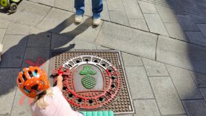 Print the City Streetart Intervention durch ErwaOne in Augsburg in Kooperation mit den Kunstsammlungen und museen von Augsburg vor dem Maximiliansmuseum (Fuggerplatz). Ein kleines Mädchen walzt Farbe auf einen Gulli-Deckel. Eine Mutter steht vor dem Kind.