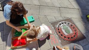 Print the City Streetart Intervention durch ErwaOne in Augsburg in Kooperation mit den Kunstsammlungen und museen von Augsburg vor dem Maximiliansmuseum (Fuggerplatz). Eine Familie taucht eine Farbwalze in Farbe um sie anschließend auf einen Gulli Deckel anzubringen.