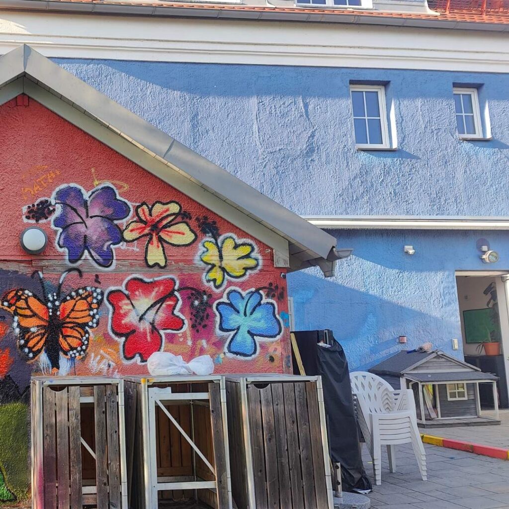 Graffiti- und Streetart Workshop mit Erwa.One in Herrsching am Ammersee. Eine von Jugendlichen bunt gestaltete Hauswand mit Blumen. Dahinter das blaue Haus Stellwerk Haus der Jugend Herrsching