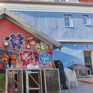 Graffiti- und Streetart Workshop mit Erwa.One in Herrsching am Ammersee. Eine von Jugendlichen bunt gestaltete Hauswand mit Blumen. Dahinter das blaue Haus Stellwerk Haus der Jugend Herrsching