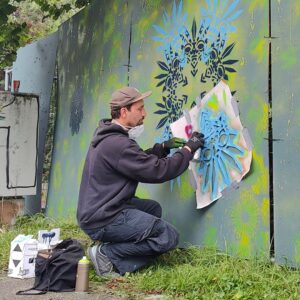 Peace Tower Jam am zukünftigen Studentenwohnheim Lechbrücke in Augsburg. Streetart und Graffiti auf Bauzäunen sorgen für Farbe und Miteinander. Der Künstler bei der Arbeit. Er sprüht eine Schablone auf die Holzwand, welche im Anschluss eine aufwendige Geometrie erzeugt.
