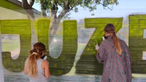 Streetart- und Graffiti Workshop im Rahmen des Ferienprogrammes des Jugendzentrums von Kirchheim an der Grund- und Mittelschule. Zwei Mädchen sprühen Buchstaben in grün auf eine mobile Folie