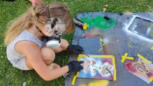 Streetart- und Graffiti Workshop im Rahmen des Ferienprogrammes des Jugendzentrums von Kirchheim an der Grund- und Mittelschule. Ein Mädchen sprüht eine Schablone auf einen bunt bemalten Karton. Eine Vogel-Schablone liegt daneben auf eienr LKW Plane