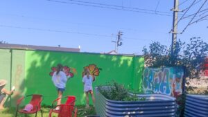 Graffiti- und Streetart Workshop mit Erwa.One in Herrsching am Ammersee. Zwei junge Mädchen sprühen auf einer grün grundierten Wand Blumen-Motive. Rechts ist ein Graffito mit dem Schriftzug Erwa zu sehen.