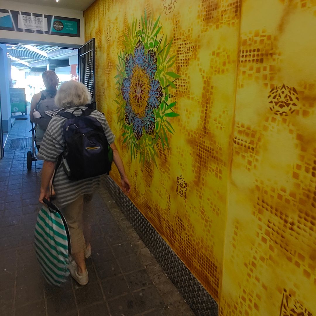 Neue Ornamente by Erwa.one Passagengestaltung der Zederpassage am Hauptplatz von Landsberg am Lech, Streetart und urbane Kunst, professionnelle Auftragsarbeit mit einzigartigen Schablonen-Motiven. Zwei Passanten laufen in die Zedergalerie an einem Ornament vorbei. Die Wand ist farbenfroh gestaltet