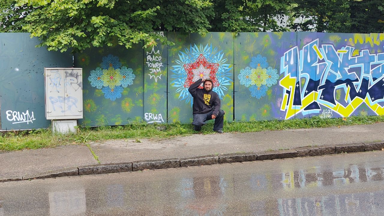 Peace Tower Jam am zukünftigen Studentenwohnheim Lechbrücke in Augsburg. Streetart und Graffiti auf Bauzäunen sorgen für Farbe und Miteinander. Das Kunst Konzept neue Ornamente ist auf dem Bild in blau und gelb zu sehen und filigranen Schablonen. Im Vordergrund der Streetartkünstler ErwaOne
