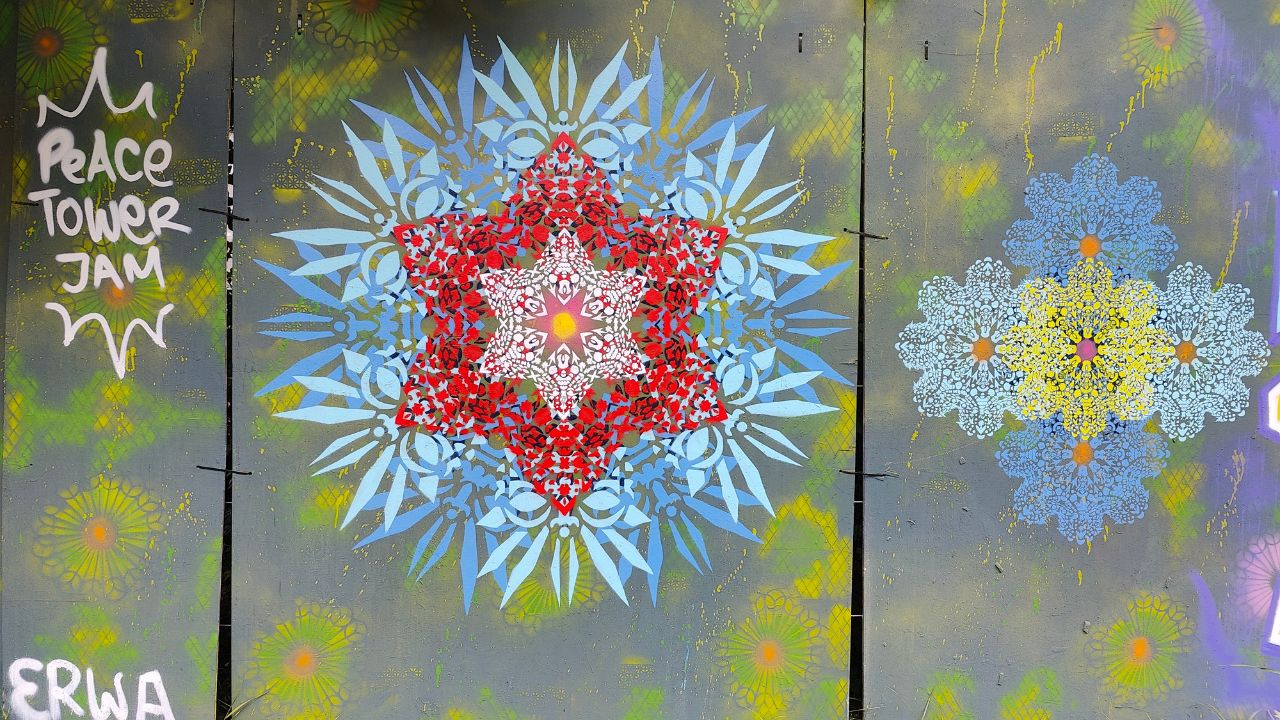 Peace Tower Jam am zukünftigen Studentenwohnheim Lechbrücke in Augsburg. Streetart und Graffiti auf Bauzäunen sorgen für Farbe und Miteinander. Das Kunst Konzept neue Ornamente ist auf dem Bild in blau und rot zu sehen und filigranen Schablonen. In der mitte ein roter Stern umgeben von blauen blüte-ähnlichen Motiven