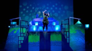 Theaterstück Jesus liebt mich Landsberg am lech. Eine Frau steht auf einer mit blau licht beleuchteten Bühnen und hält die Hände erhoben.