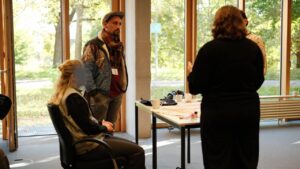 Teilnahme des Künstlers ErwaOne am Workshop Mlab Taschen mit Fragen bemalen. Der künstler steht und hat die hände in der hosentasche. Eine Person sitzt und eine Person steht. ein Tisch befindet sich zwischen der Menschengruppe. Die Veranstaltung fand im Zentrum für Klimaresilienz der Universität Augsburg statt