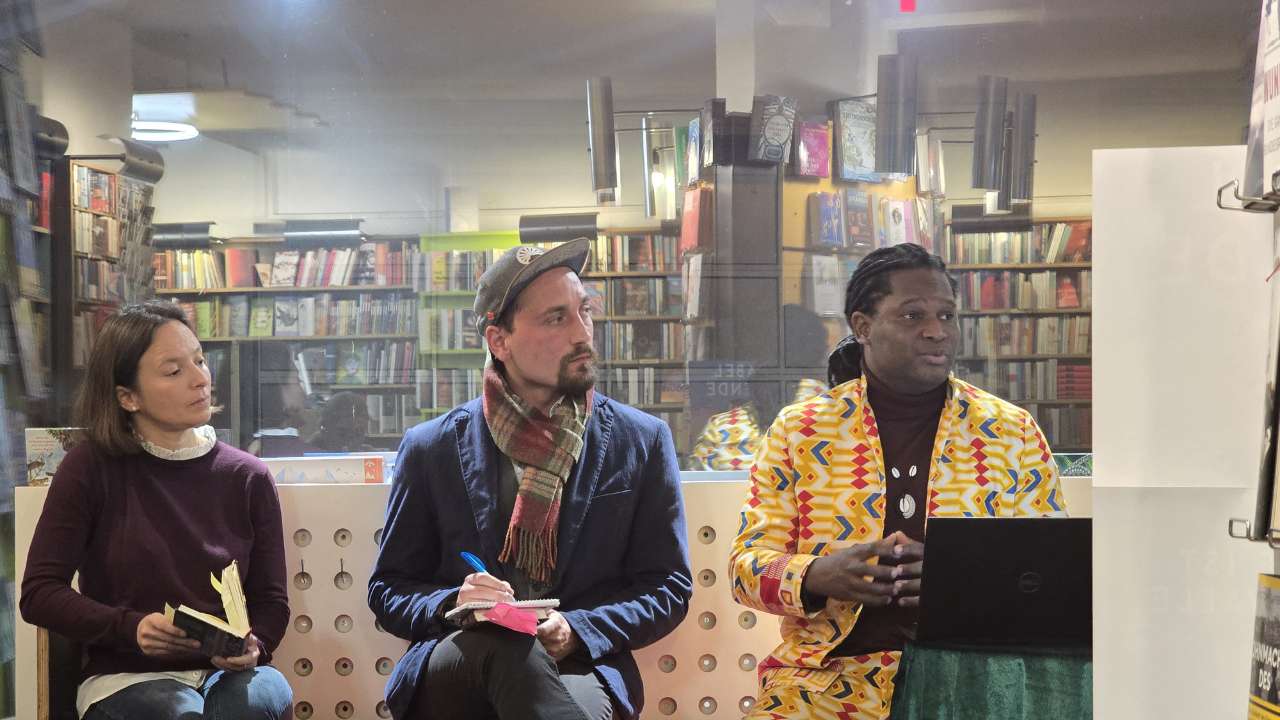 Lesung Buch Cave72 afrikanischer Autor Fann Attiki aus Brazzaville, ErwaOne als deutsch-französischer Dolmetscher, links Julia Kabatas von der solidarischen Werkstatt AUgsburg, in der Mitte Erwa.One welcher Gesprächsnotizen aufnimmt und rechts der Autor Fann Attiki welcher aus seinem Buch erzählt