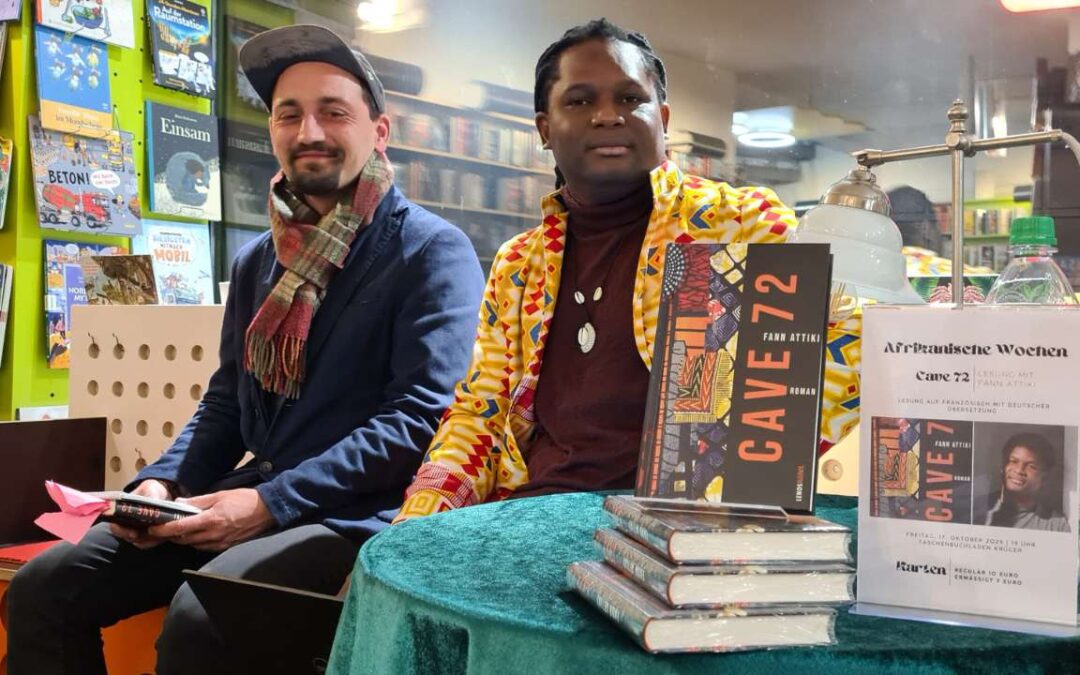 Lesung Buch Cave72 afrikanischer Autor Fann Attiki aus Brazzaville, ErwaOne als deutsch-französischer Dolmetscher, Beide Künstler sitzen auf einer Bank im Taschenbuchladen. Vor ihnen steht ein Tisch mit dem aktuellen Exemplar von Fann Attiki