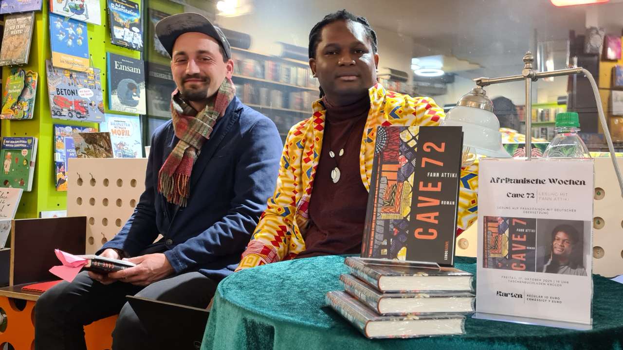Lesung Buch Cave72 afrikanischer Autor Fann Attiki aus Brazzaville, ErwaOne als deutsch-französischer Dolmetscher, Beide Künstler sitzen auf einer Bank im Taschenbuchladen. Vor ihnen steht ein Tisch mit dem aktuellen Exemplar von Fann Attiki