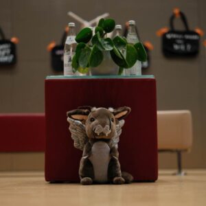 Ein Wolpertinger steht vor einem roten Sitzwürfel auf welchem eine Zimmerpflanze und Wasserflaschen stehen