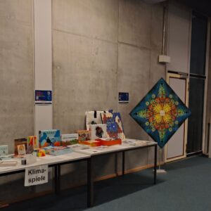 Wissenschaftliche Tagung Neue Kulturgeographie 2025 in Augsburg zum Thema "Speculative geographies of the new climate regime". Ausstellungsraum im Rahmen der wissenschaftlichen Konferenz kritischer Geographen mit links einer Auswahl an Serious Games, sowie rechts einem Kunstwerk auf Leinwand namens "Sternrose" vom Streetart und Urban-Art Künstler Erwa.One.