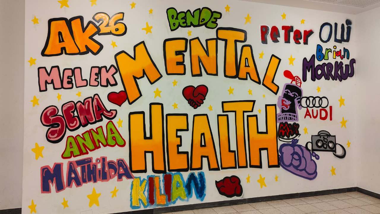 Kunstprojekt Mental Health Coach an Schulen. Graffiti-Wandbild im Keller des Förderzentrums entstanden mit Schülern und dem Künstler Erwa.One. Der Schriftzug Mental Health steht in großen organgenen Farbtönen im Zentrum der Wand. Darum herum Namen der Teilnehmer in verschiedenen Farben, sowie gelbe Sterne als Verbindendes Element.