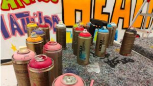 Sprühdosen stehen auf einem Tisch. Im Hintergrund ist das unfertige Wadbild zu sehen mit bunt bespühten Flächen und Buchstaben. Die Dosenfarben magenta pink orange violett rt und türkus, sowie schwarz und weiß stehen auf dem Tisch.