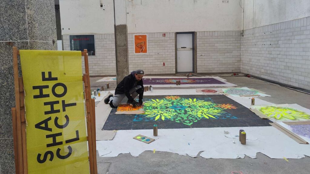 Streetart Künstler Erwa.One bei der Vorbereitung eines riesigen Kunstwerkes mit seinem Designkonzept Neue Ornamente für die Pforzheimer Kulturnacht. Der Künstler kniet in einem ehemaligen Schlachthof auf dem Boden und besprüht eine Plane.