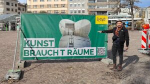 Waisenhausplatz Pforzheim Künstler ErwaOne zeigt auf einen Baubanner mit dem Transparent "Kunst braucht Räume" Sonniger Herbsttag Stadtzentrum