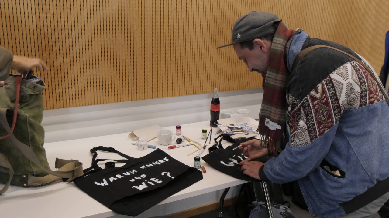 Mlab Workshop im Zentrum für Klimaresilienz der Universität Augsburg. Taschen mit Fragen bemalen, um diese in die Welt zu tragen. Der Künstler Erwa.one bemalt eine Tasche mit weißer Farbe. Er trägt eine blaue Recycling Jacke, einen Schal und eine Cap. Auf dem Tisch liegt eine weitere (schwarze) Tasche mit der Frage "Warum anders und wie?"