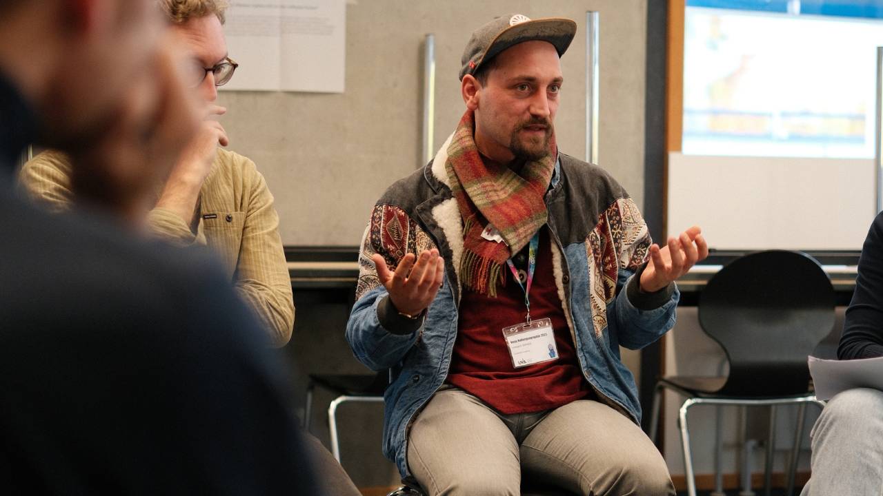 Künstler Erwa.One während seines Redebeitrags zum Thema "Streetart als Change Agent im neuen Klimaregime" im Workshop zum Thema geographische Bildung für planetare Zukünfte. Er hält die Hände vom Körper weg. Blaue Jacke und Cap auf dem Kopf mit einem Schal