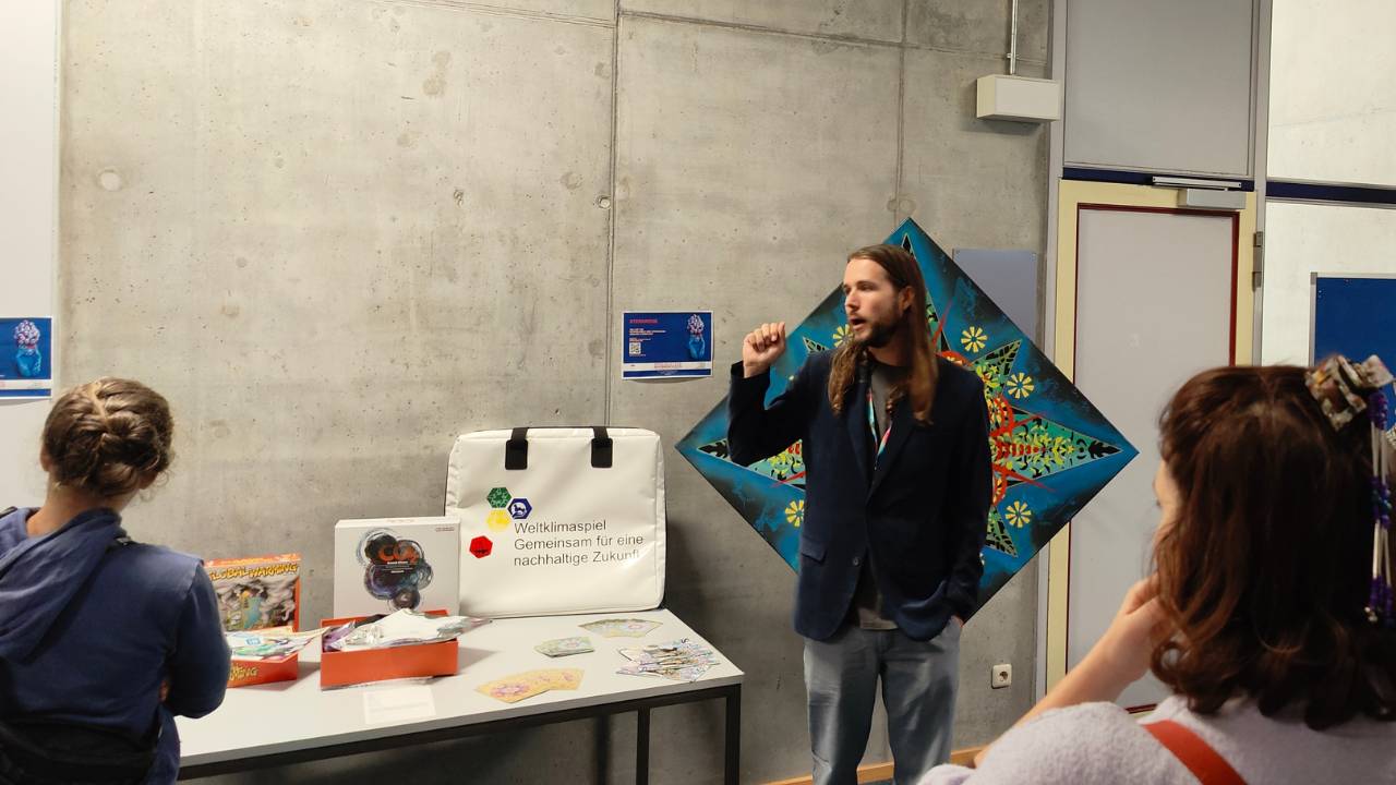 Wissenschaftliche Tagung Neue Kulturgeographie 2025 in Augsburg zum Thema "Speculative geographies of the new climate regime". Rouven Kaiser erklärt die Ausstellung im Rahmen der Konferenz und zeigt auf eine Auswahl an Serious Games - Spiele um den Klimawandel zu verstehen. Hinter ihm ist ein Kunstwerk von Erwa.One zu sehen, die "Sternrose", welche er 2022 im Atelier Rose von Schondorf gestaltet hat.