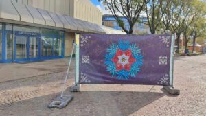 ErwaOne Pforzheimer Kulturnacht Kunst im öffentlichen Raum mit Werkpräsentation von besprühten Banner mit den "Neuen Ornamenten". Auf dem Bild in Pforzheim-Waisnehausplatz Stadtzentrum zu sehen, gegenüber vom Stadttheater. Blau Rotes Ornament mit violettem Hintergrund.
