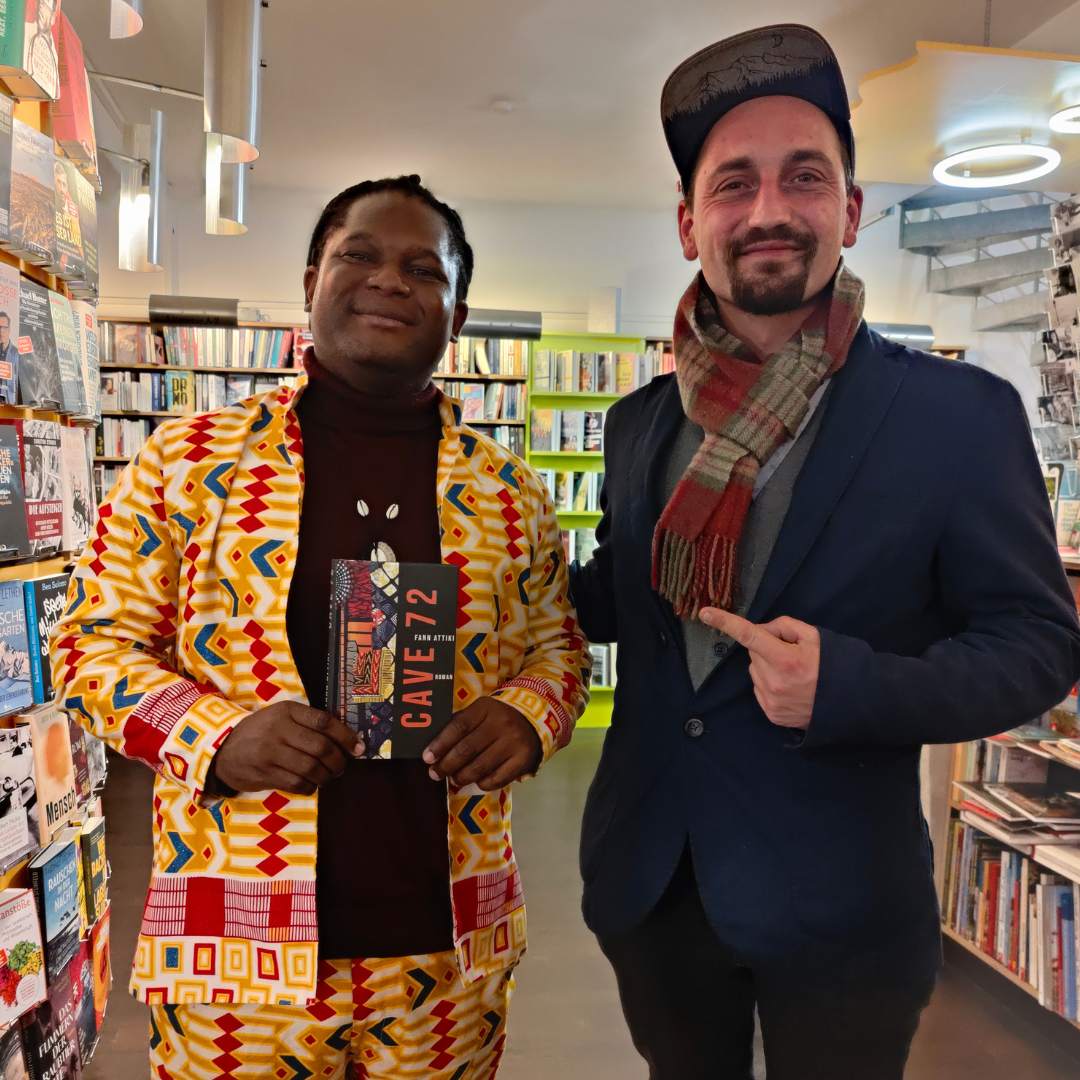 Lesung Buch Cave72 afrikanischer Autor Fann Attiki aus Brazzaville, ErwaOne als deutsch-französischer Dolmetscher, beide Künstler stehen in einem Buchladen. Fann hält ein Buchexemplar in die Kamera, ErwaOne zeigt mit einem Finger auf Ihn.